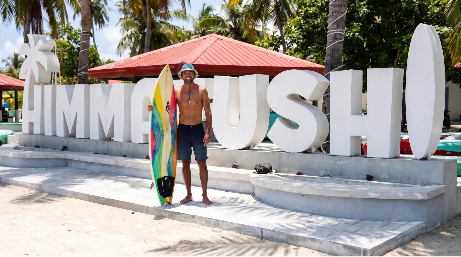 Standing with a surfboard at the Himmafushi beach sign in the Maldives